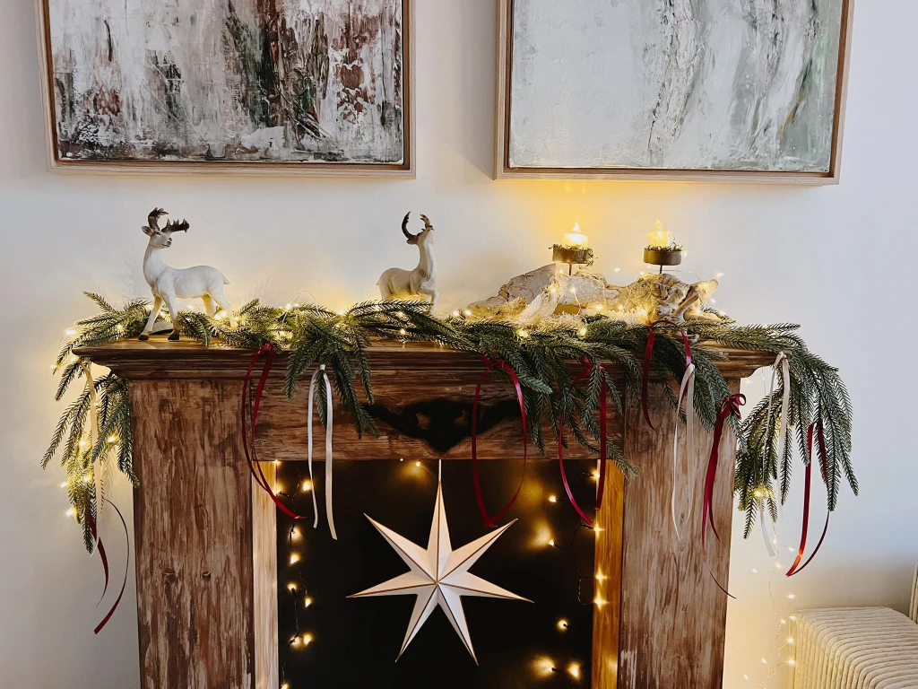 Kominek ozdobiony świątecznym stroikiem z gałązek, światełkami LED oraz figurkami reniferów i stojącą gwiazdą, na tle dwóch abstrakcyjnych obrazów.