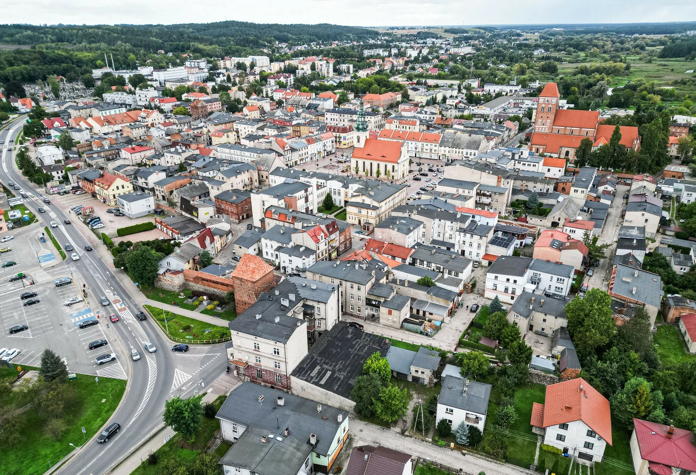 To najbiedniejsze miasto w Polsce. Lokalizacja może być zaskoczeniem Panorama niewielkiego miasta z licznymi budynkami o czerwonych i szarych dachach, wyraźnie widocznym dużym kościołem oraz placem z samochodami po lewej stronie, otoczonym zielenią oraz pagórkami w tle.