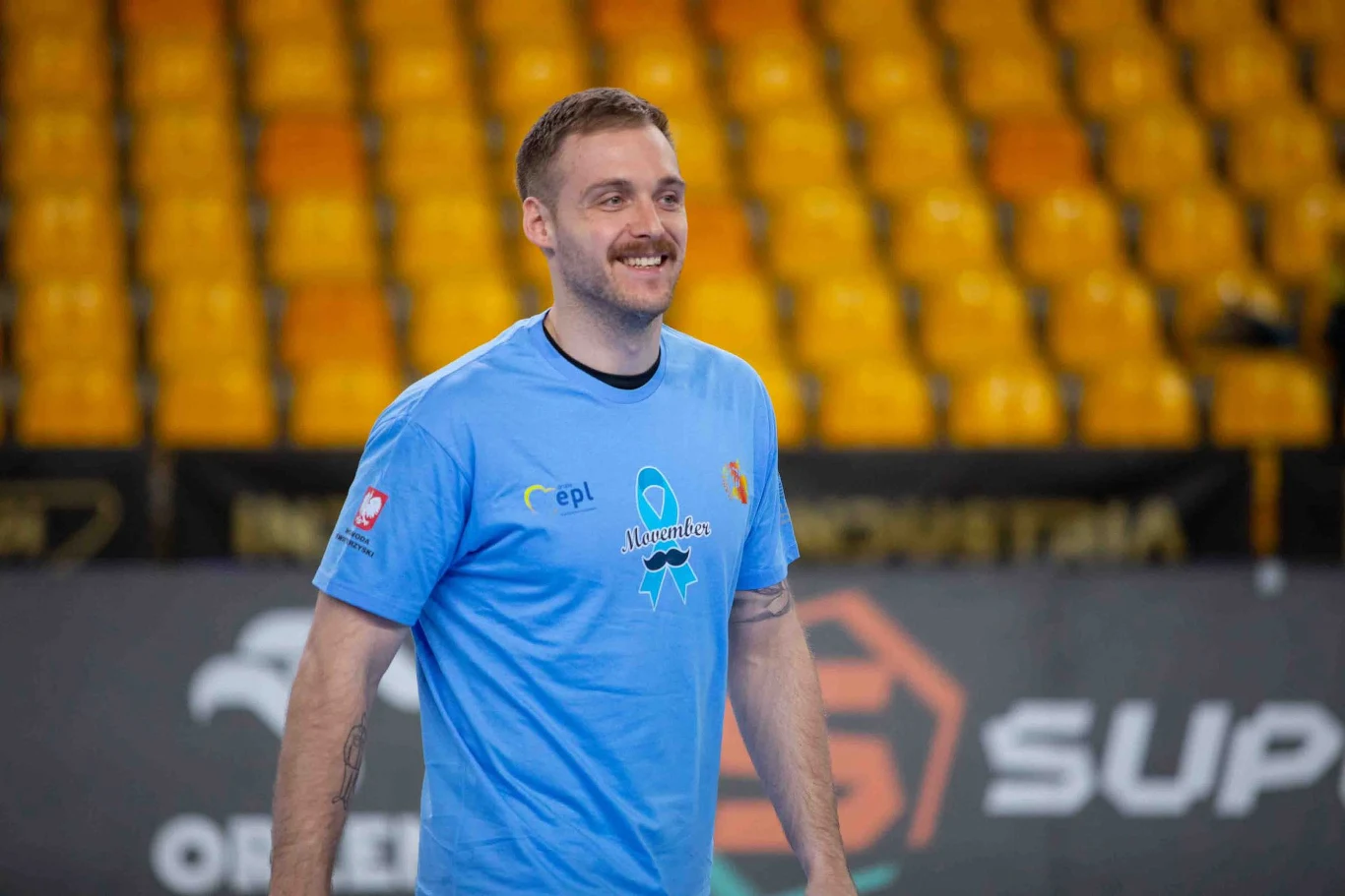 Zakończyła się akcja "Movember" Uśmiechnięty mężczyzna w jasnoniebieskiej koszulce sportowej stoi na tle pustych żółtych krzeseł na hali sportowej.