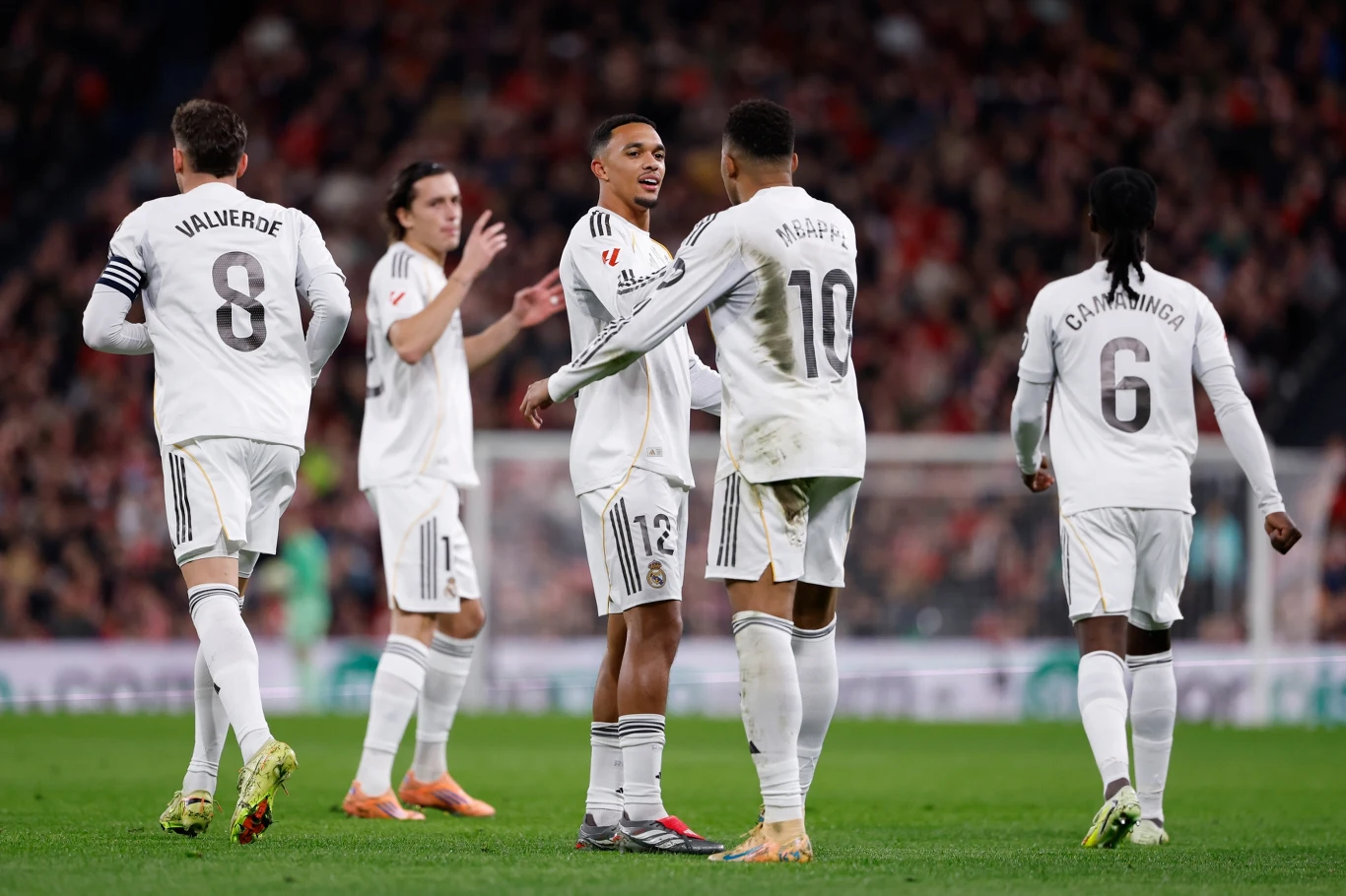 Piłkarze Realu Madryt Zawodnicy drużyny piłkarskiej ubrani w białe stroje celebrują udaną akcję podczas meczu. W tle widoczny stadion oraz publiczność na trybunach.