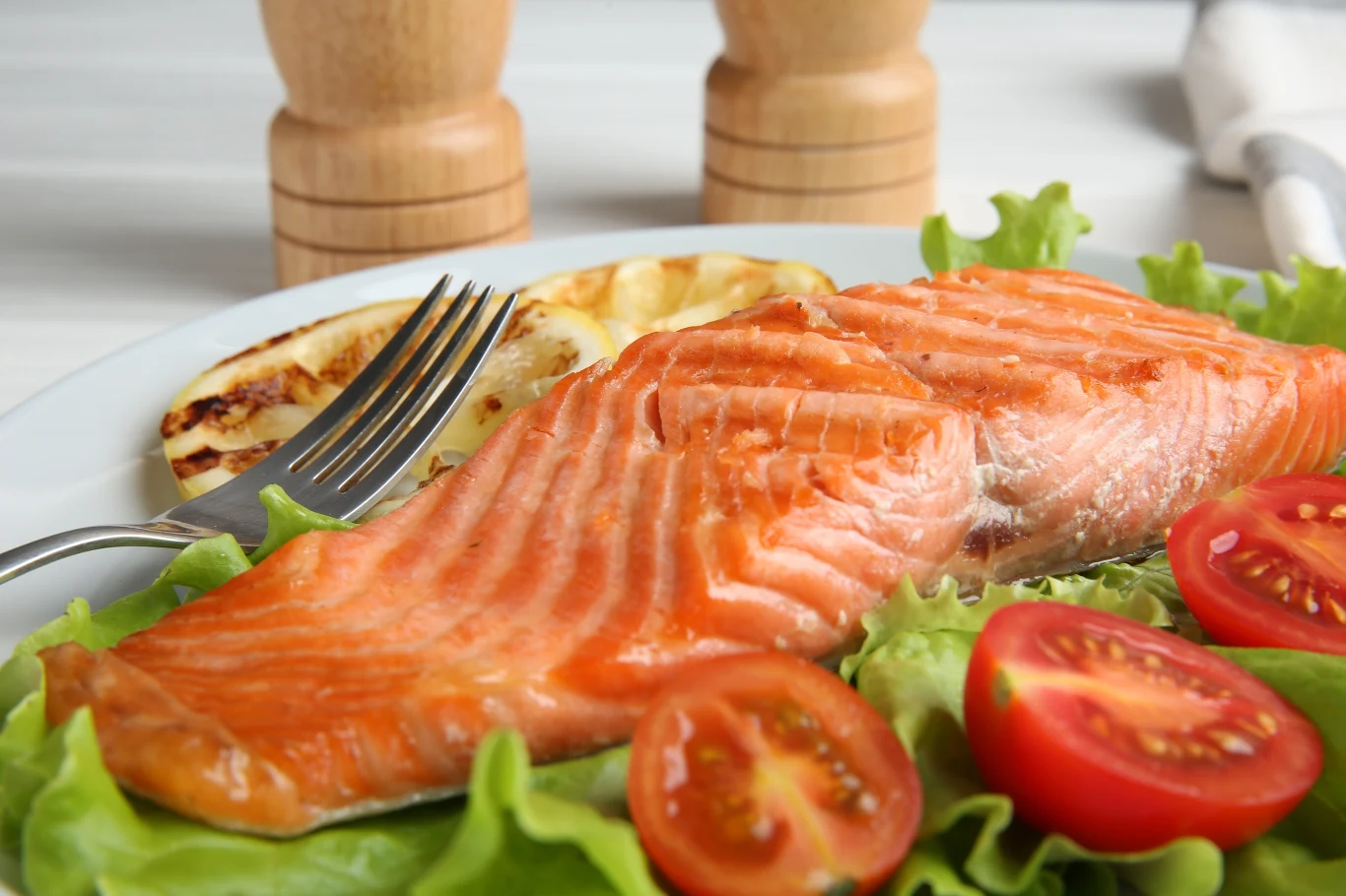 Porcja pieczonego łososia ułożona na liściach sałaty, pokrojone pomidorki koktajlowe oraz plaster grillowanej cytryny, w tle dwa młynki do przypraw.