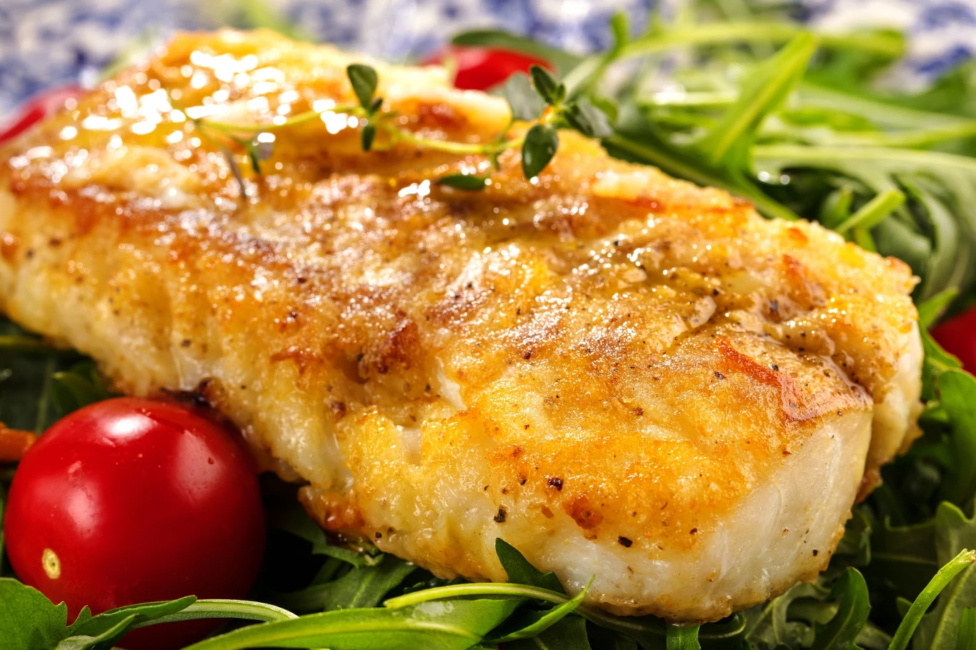 Soczysty kawałek pieczonej ryby na liściach świeżej rukoli, obok kilka pomidorków koktajlowych; całość udekorowana gałązką tymianku, danie apetyczne i starannie przygotowane.