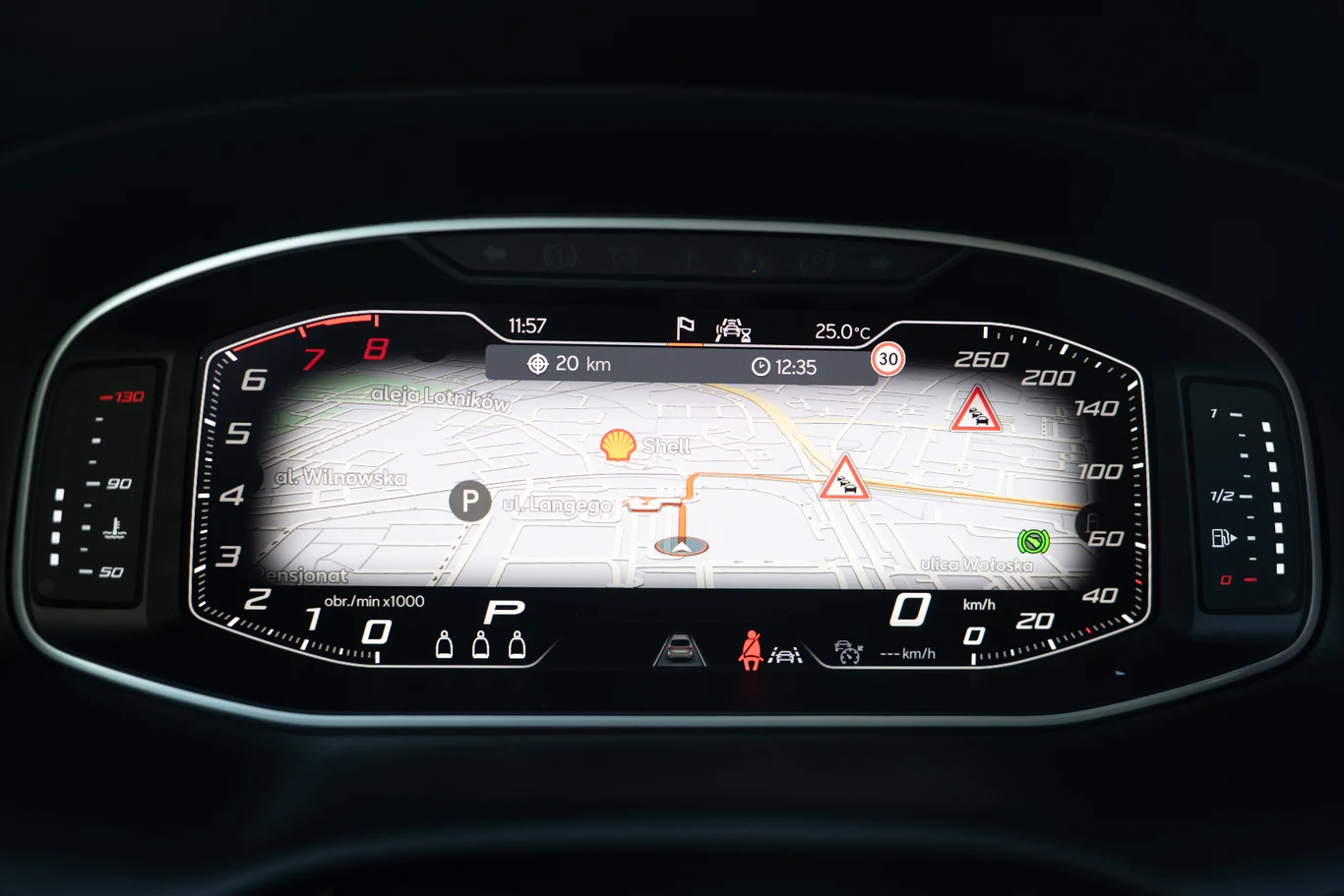 Cyfrowy kokpit samochodu z wyświetlanym widokiem nawigacji GPS, licznikiem prędkości, obrotomierzem oraz oznaczeniami ostrzegawczymi i informacjami o trasie.