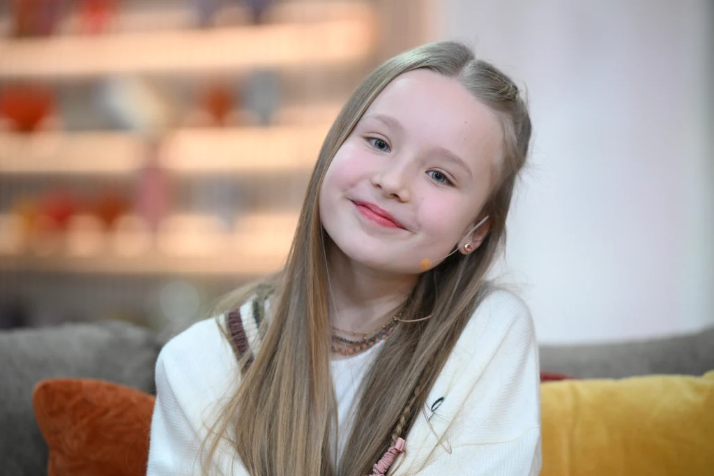 Uśmiechnięta dziewczynka z długimi blond włosami, ubrana w jasny sweter, siedzi na kanapie na tle rozmytego wnętrza z kolorowymi półkami. Jej postawa jest zrelaksowana, a twarz łagodna.