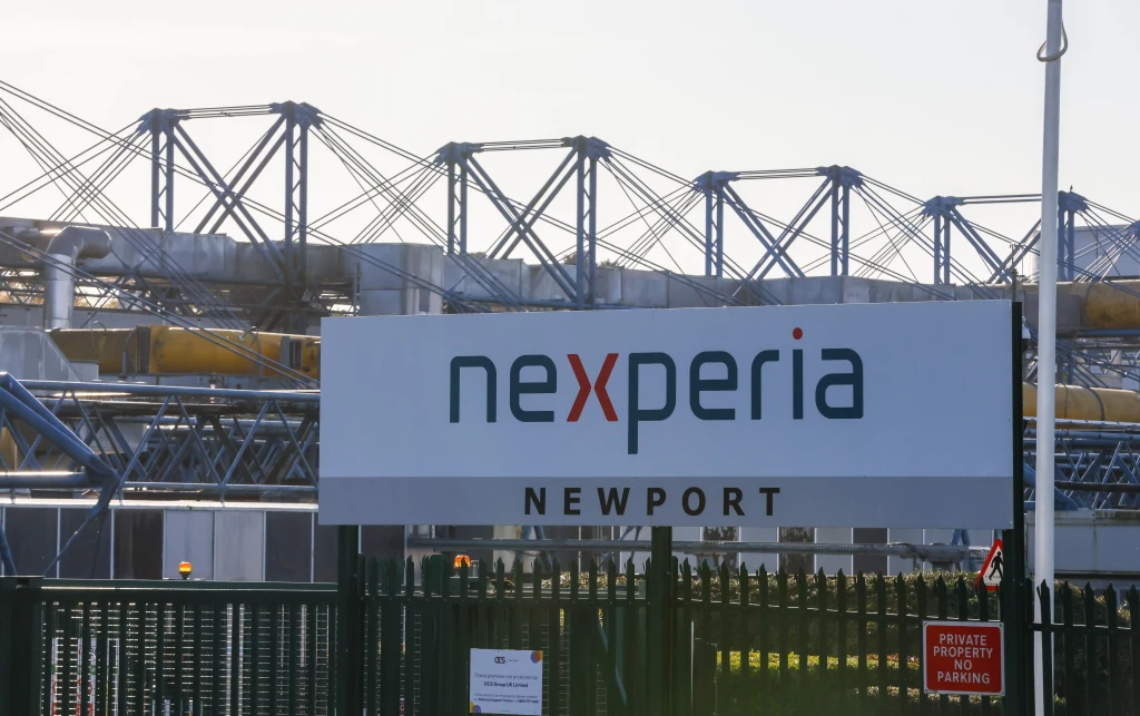 To jeszcze nie koniec sporu Chin i Holandii o Nexperię. | Fot. ilustracyjna Tablica z logo firmy Nexperia i napisem Newport znajdująca się przed ogrodzeniem przemysłowego kompleksu. W tle konstrukcje metalowe i elementy budynków zakładowych.