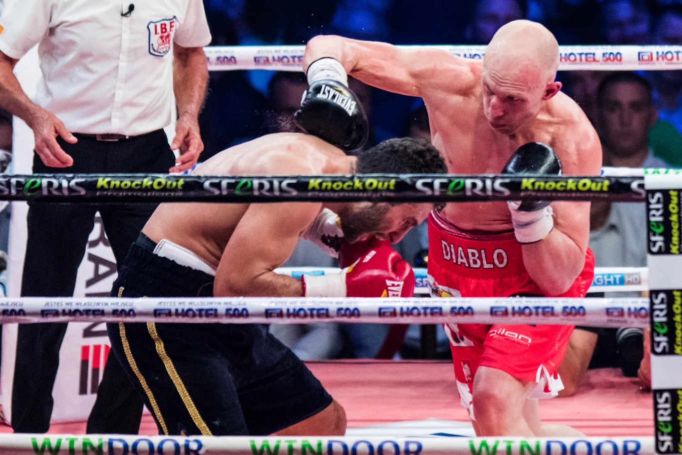 Dwaj bokserzy walczą w ringu, jeden z nich zadaje cios przeciwnikowi w pochylonej pozycji, wokół widoczne są bandy reklamowe i sędzia obserwujący pojedynek.