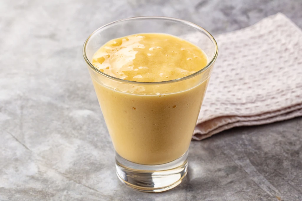 Szklanka kremowego koktajlu o jasnożółtej barwie stoi na jasnym, marmurowym blacie. W tle widoczna jest ułożona ściereczka w delikatnych kolorach.
