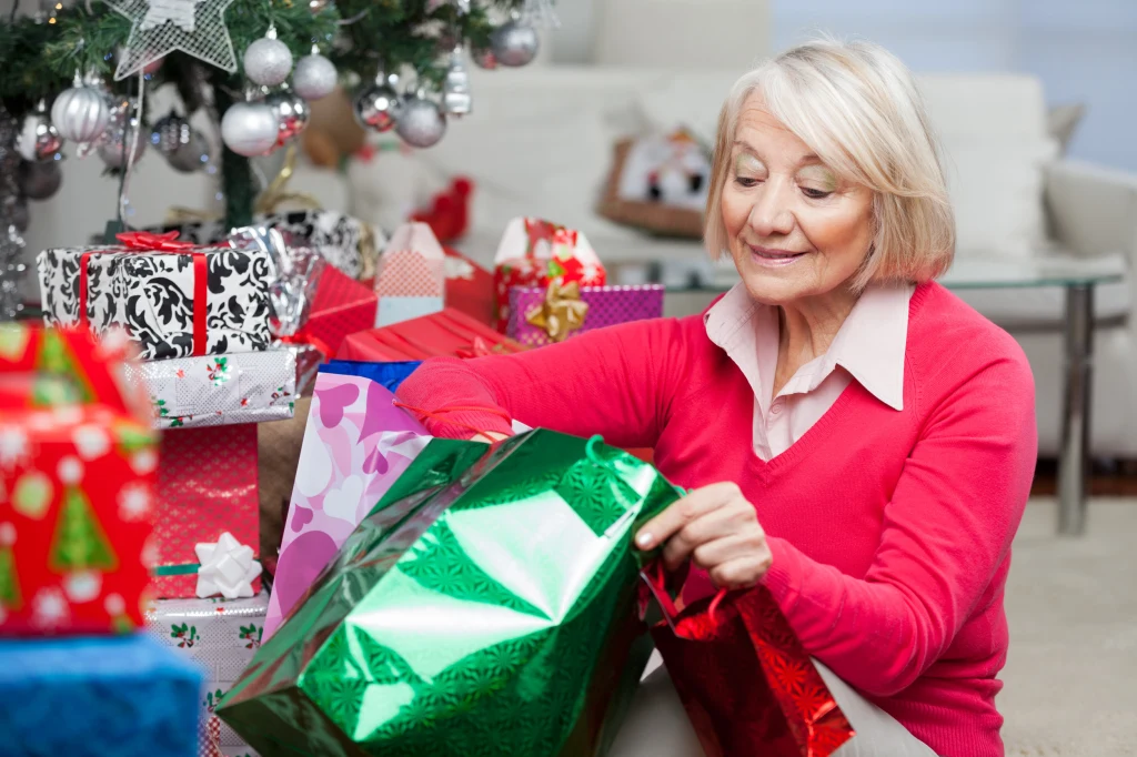 Starsza kobieta siedzi przy choince, rozpakowuje kolorowe prezenty, wokół niej widać wiele opakowanych paczek świątecznych i świąteczne dekoracje.