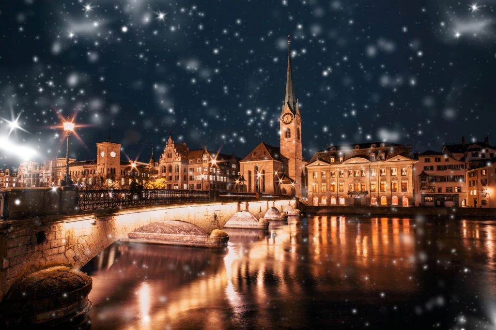 Most oświetlony żółtym światłem, odbijającym się w wodzie rzeki, zabytkowy kościół z wysoką wieżą na tle nocnego nieba oraz śnieg padający na urokliwe, historyczne centrum miasta.