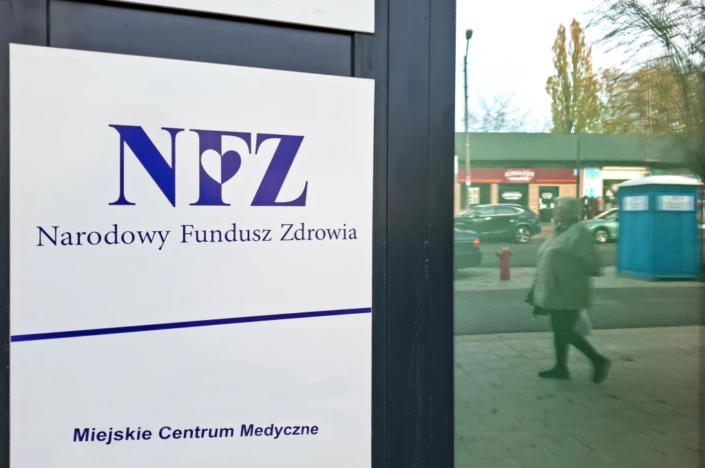 Tablica z logotypem Narodowego Funduszu Zdrowia oraz napisem Miejskie Centrum Medyczne, w tle odbicie spacerującej osoby, elementy miejskiej architektury i niebieska toaleta przenośna.
