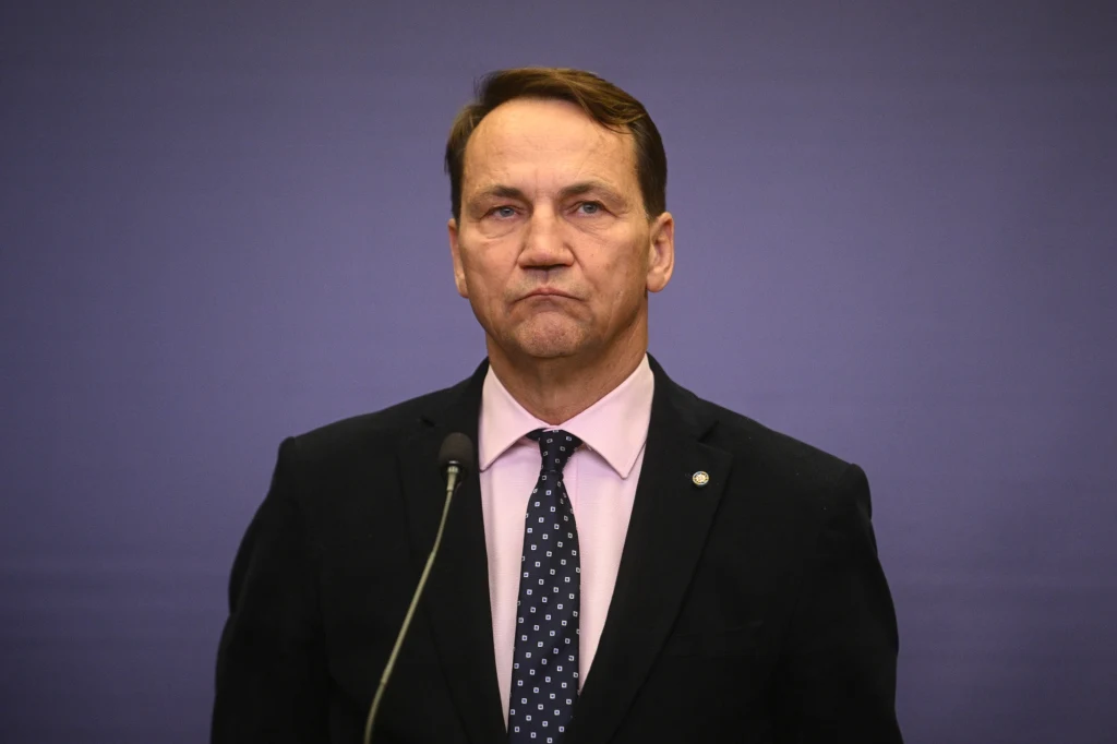 Mężczyzna w garniturze i krawacie stoi przed mikrofonem na tle jednokolorowej ściany, patrząc poważnie przed siebie.