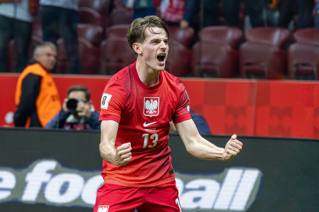Piłkarz reprezentacji Polski w czerwonym stroju z numerem 13 świętuje sukces z zaciśniętymi pięściami i okrzykiem, w tle trybuny oraz kibice.