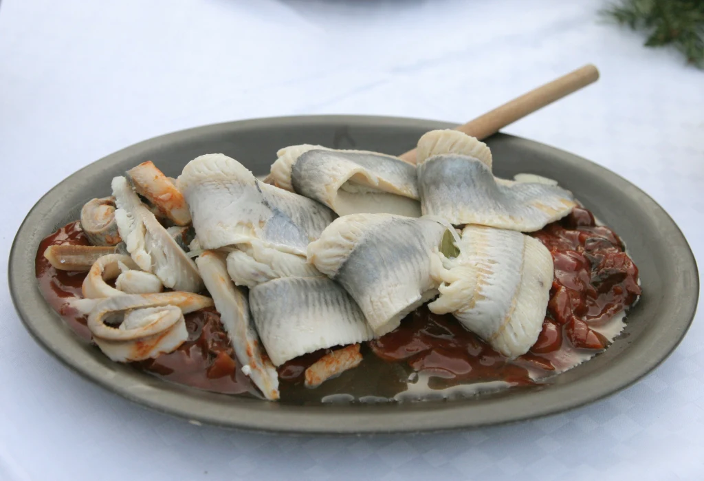 Porcja śledzi ułożona na owalnym talerzu, podanych na czerwonym sosie z warzywami, z charakterystycznie zawiniętymi filecikami rybnymi i łyżką do serwowania.