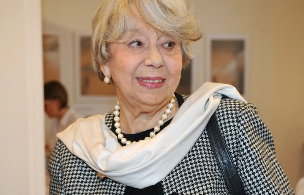 Zofia Kucówna Starsza kobieta o jasnych włosach i eleganckim wyglądzie, ubrana w kratkowaną marynarkę, biały szalik i naszyjnik z pereł, z delikatnym uśmiechem, znajduje się w pomieszczeniu o jasnych ścianach.