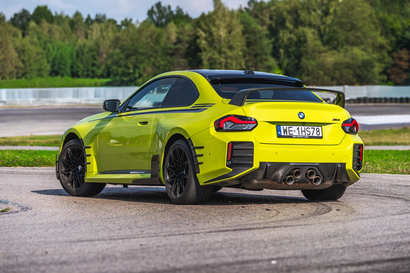 480 KM i napęd tylko na tył sprawiają, że BMW M2 potrafi mieć naprawdę agresywny charakter Jaskrawożółty sportowy samochód BMW z czarnymi akcentami, widok od tyłu na torze wyścigowym, masywne tylne skrzydło, cztery końcówki wydechu, czarne felgi, w tle drzewa i bariera ochronna.