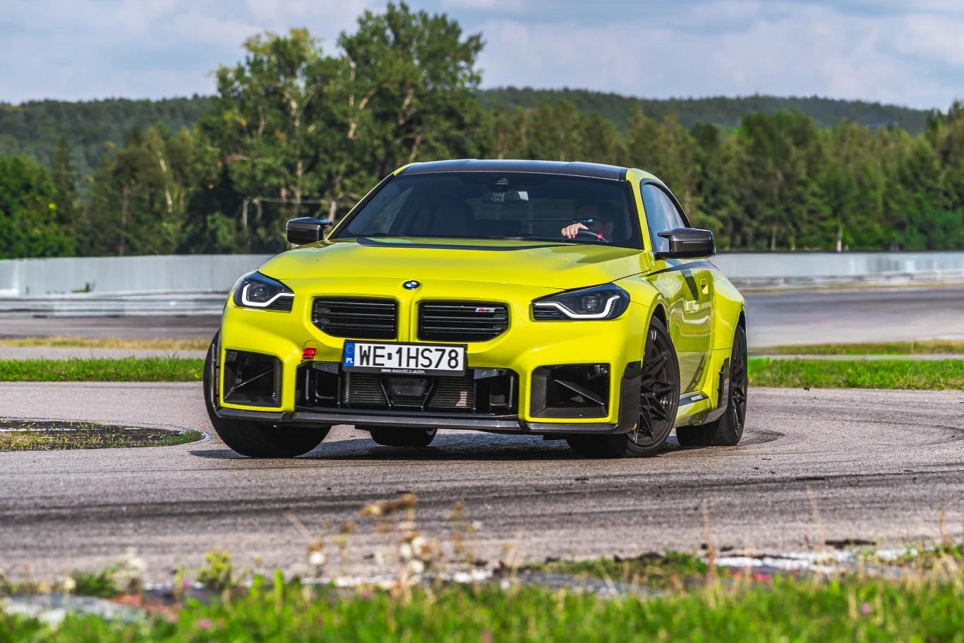 BMW M2 Jaskrawożółte sportowe BMW M2 wykonuje dynamiczny zakręt na asfaltowym torze wyścigowym, otoczone zielenią i drzewami w tle, na tablicy rejestracyjnej widoczny polski numer.