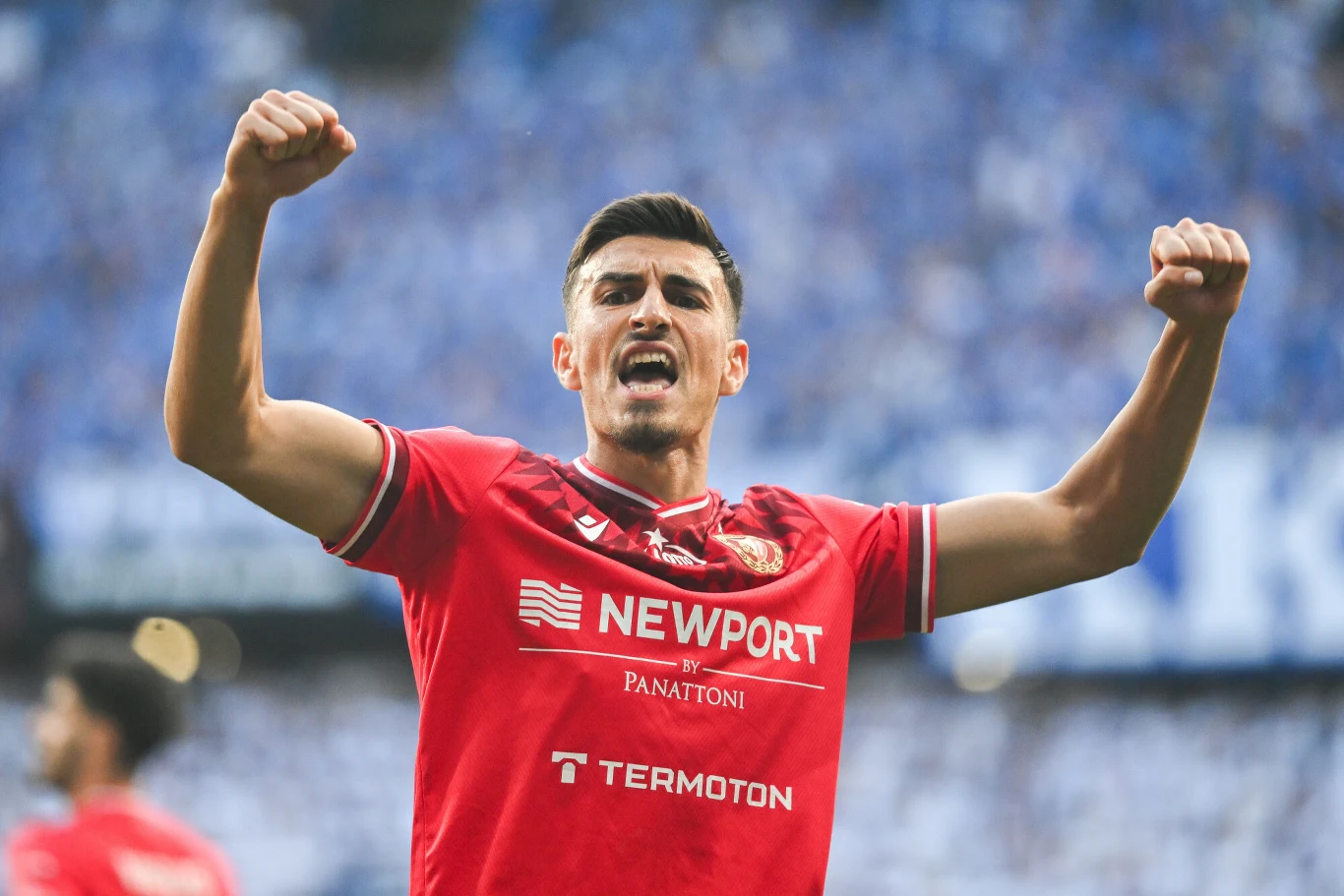 Piłkarz w czerwonej koszulce z uniesionymi w górę rękami wyraża radość po zdobyciu gola, w tle rozmazana widownia stadionu.