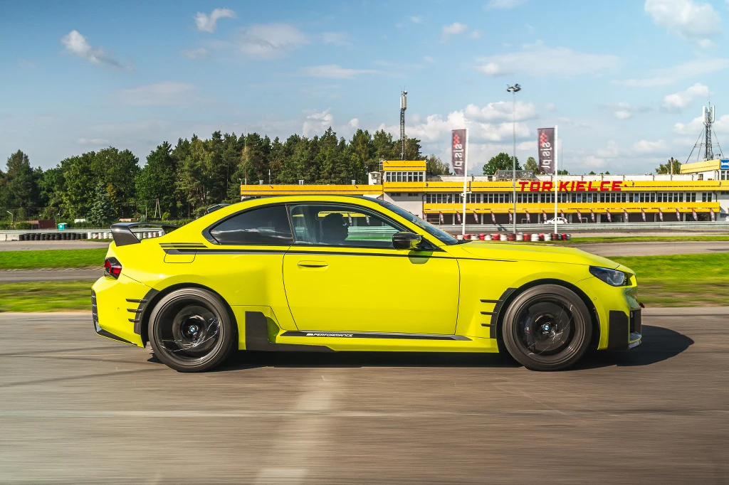 Jaskrawożółty sportowy samochód pędzący po torze wyścigowym, w tle obiekt sportowy z napisem Tor Kielce, otoczony zielenią oraz trybunami.