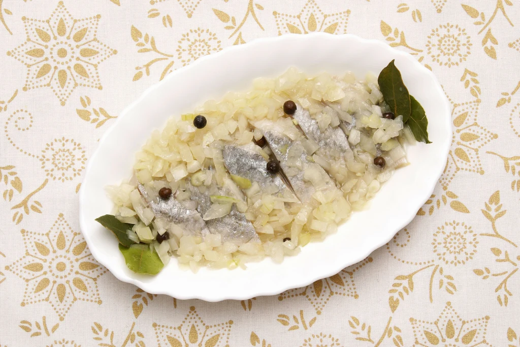 Kawałki śledzia ułożone na białym półmisku, przykryte pokrojoną w drobną kostkę cebulą oraz przyprawione liśćmi laurowymi i ziarnami czarnego pieprzu, całość podana na ozdobnym obrusie.