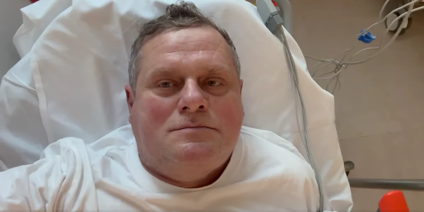 Kazik Staszewski trafił do szpitala Mężczyzna w średnim wieku leży na białej szpitalnej pościeli, ubrany w białą koszulkę, z widocznymi rurkami medycznymi w tle po prawej stronie.