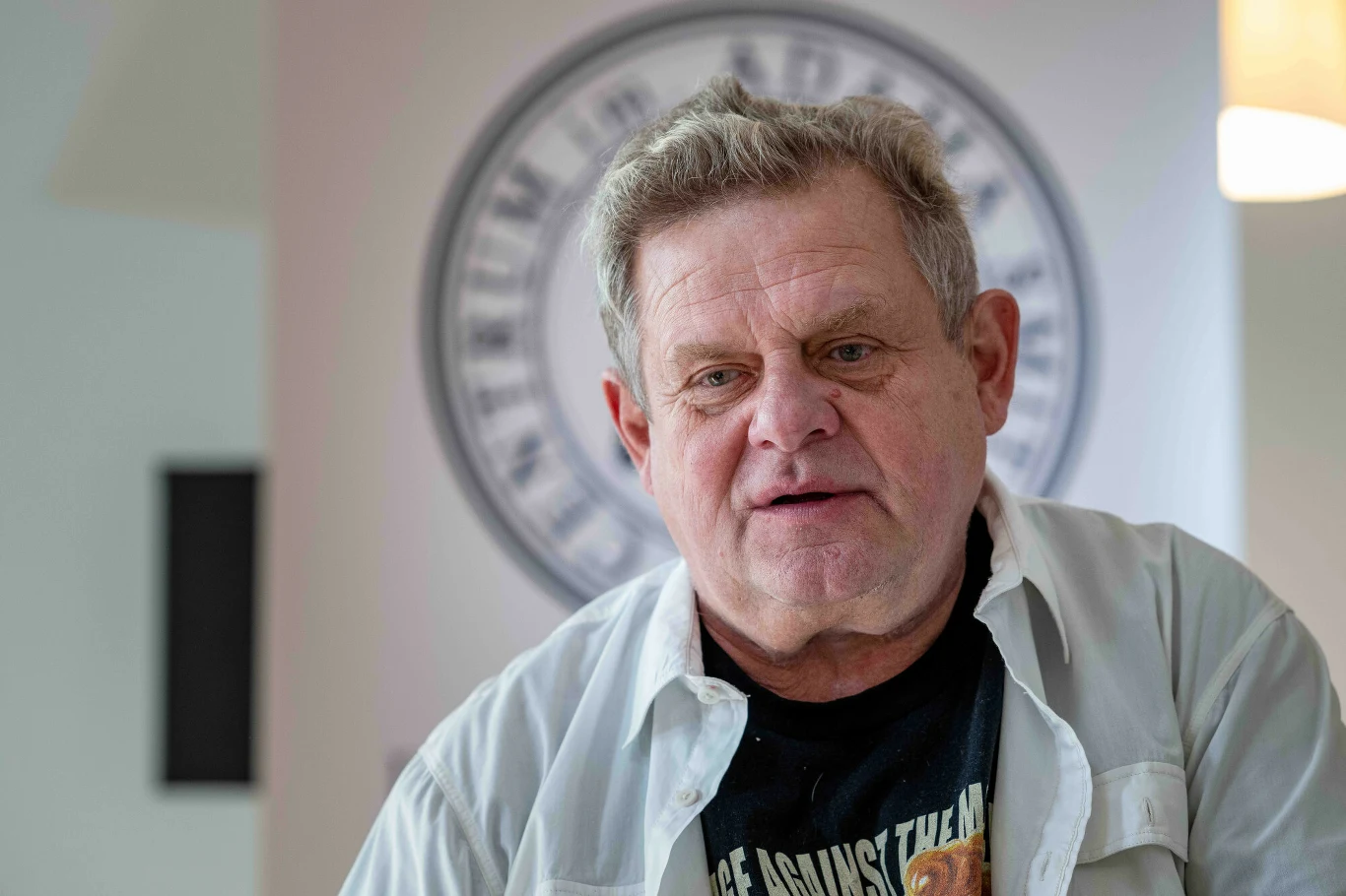 Kazik Staszewski trafił do szpitala Starszy mężczyzna z siwymi włosami ujęty w zbliżeniu, ubrany w jasną koszulę oraz ciemny t-shirt, w tle rozmyte logo na okrągłej tablicy.