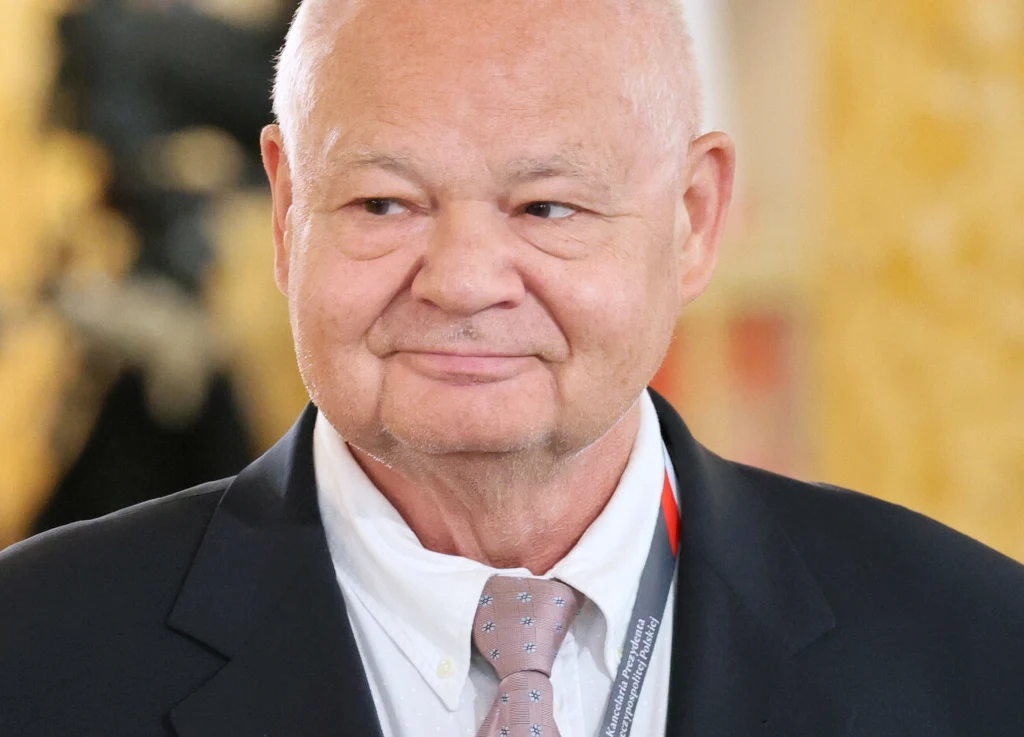 Jakie będą dalsze ruchy RPP? Adam Glapiński uważa, że Rada będzie chciała przejść w tryb "wait and see" Starszy mężczyzna w garniturze z jasną koszulą i krawatem, lekko uśmiechnięty, w tle rozmyte żółte i czarne elementy, wyraźny portret z naciskiem na twarz.