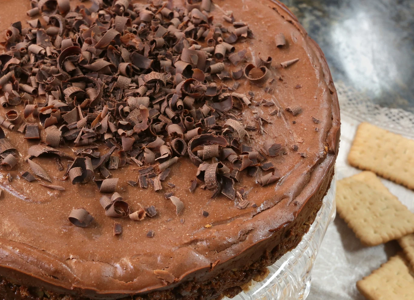 Sernik czekoladowy w mig zniknie ze stołu Czekoladowe ciasto udekorowane czekoladowymi wiórkami na wierzchu, ustawione na szklanej paterze, obok leżą jasne herbatniki.