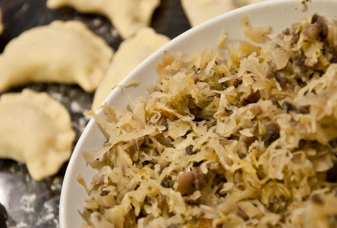 Świąteczna kapusta na wiele sposobów: albo solo, albo jako farsz do pierogów Miseczka wypełniona farszem z kapusty i grzybów na pierwszym planie, w tle kilka nieugotowanych pierogów leżących na blacie.