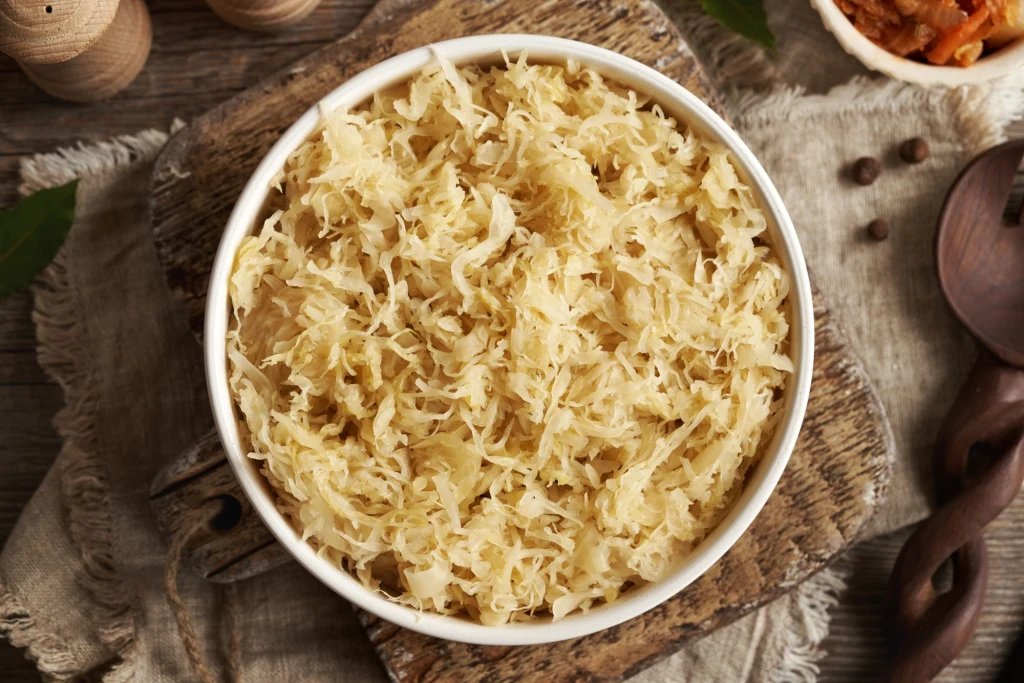 Miseczka wypełniona kiszoną kapustą ustawiona na drewnianej desce, otoczona elementami kuchennymi, przyprawami i lnianą serwetką.