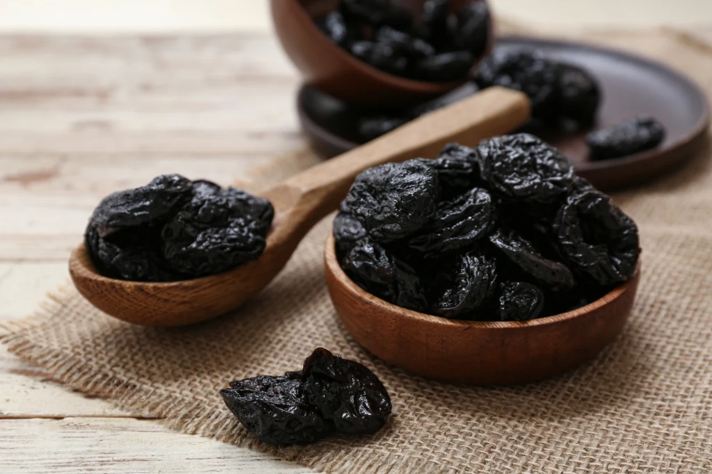 Suszone śliwki ułożone w dwóch drewnianych miseczkach oraz na drewnianej łyżce, całość ustawiona na jutowym kawałku materiału na drewnianym stole.