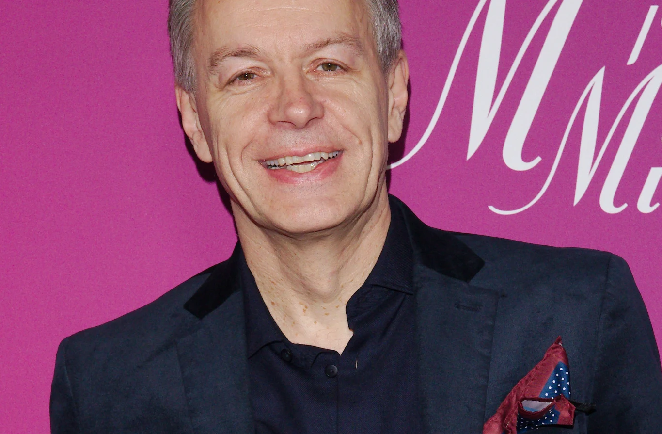 Gwiazdor "M jak miłość" wraca do serialu Uśmiechnięty mężczyzna w granatowej marynarce i czarnej koszuli, elegancko ubrany, stojący na tle różowej ścianki z białym napisem.