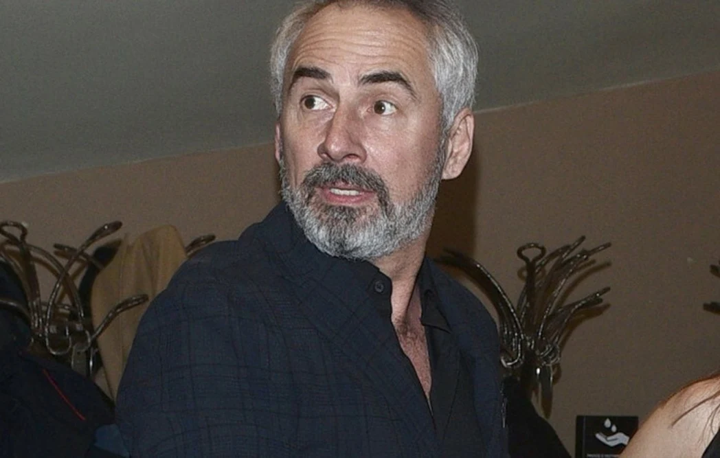 Jacek Kopczyński Mężczyzna z siwą brodą i włosami w ciemnej koszuli spogląda w bok, w tle widoczne są wieszaki z ubraniami oraz jasna ściana.