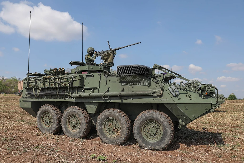 Opancerzony pojazd wojskowy na ośmiu kołach, wyposażony w wieżyczkę z zamontowanym karabinem maszynowym, na dachu znajduje się żołnierz w pełnym umundurowaniu. Pojazd stoi na otwartym, suchym terenie pod niebieskim niebem z kilkoma chmurami.