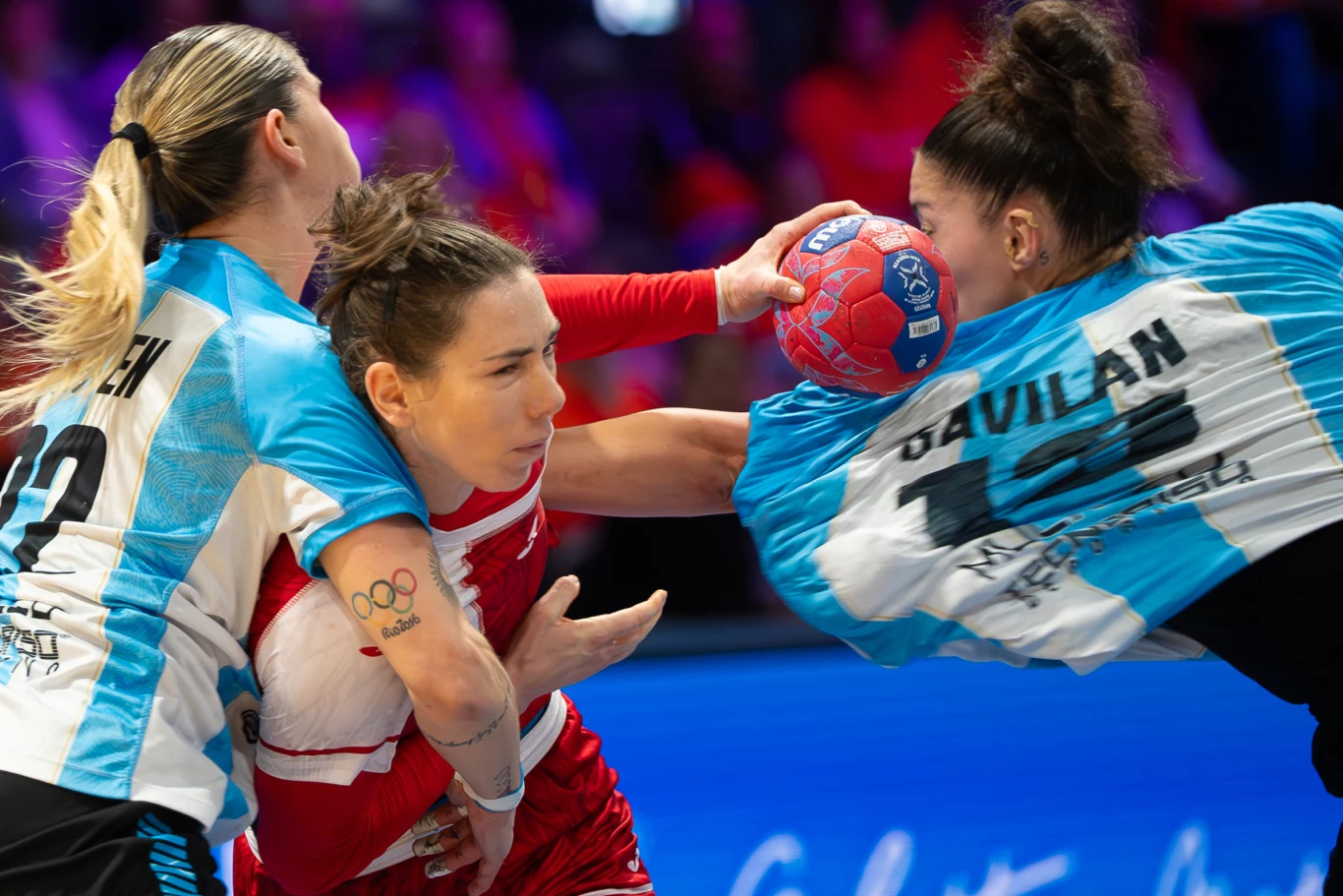 Zacięta walka o piłkę podczas meczu piłki ręcznej kobiet, dwie zawodniczki w niebiesko-białych strojach bronią się przed atakiem zawodniczki w czerwonym stroju, skupienie i dynamika ruchu widoczne na twarzach wszystkich uczestniczek.