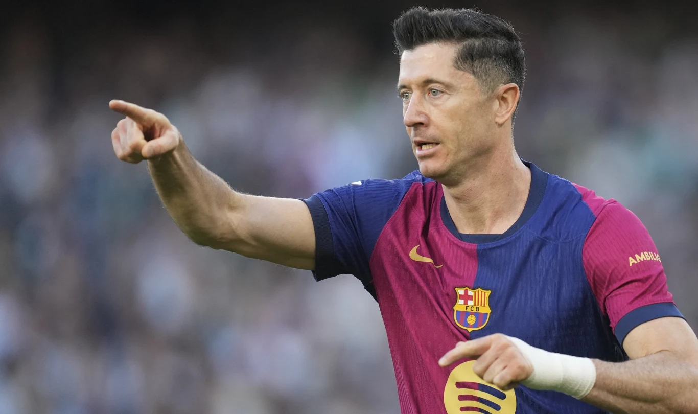Robert Lewandowski Piłkarz w stroju FC Barcelona z uniesionym ramieniem wskazuje wyciągniętym palcem kierunek, na dłoni widoczny bandaż. W tle rozmyta publiczność na stadionie.