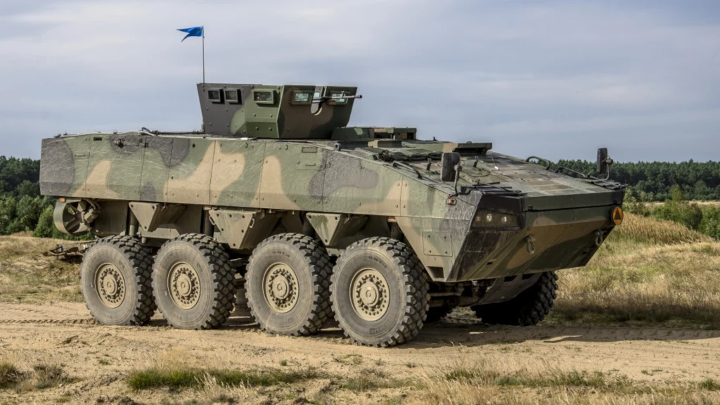 Opancerzony pojazd wojskowy na ośmiu kołach w zielonym kamuflażu znajduje się na piaszczystym terenie w otwartej przestrzeni, w tle las i niebo z lekkim zachmurzeniem, na pojeździe widoczna niewielka niebieska flaga.