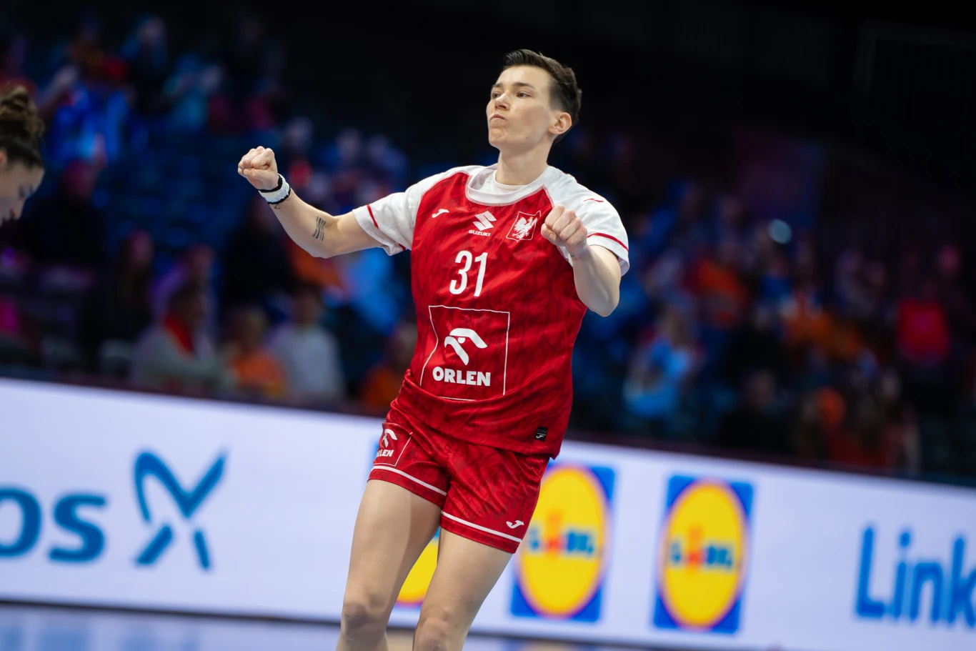 Zawodniczka w czerwonym stroju sportowym z numerem 31 na koszulce, w trakcie emocjonującego meczu piłki ręcznej na hali, z widocznym logo sponsora i tłumem kibiców w tle.