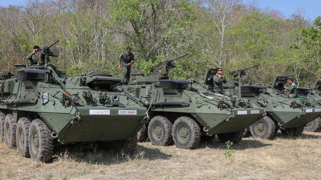 Cztery wojskowe pojazdy opancerzone z żołnierzami na pokładzie, ustawione w szeregu na trawiastej polanie, w tle widoczne drzewa i zarośla. Żołnierze wyposażeni w hełmy i karabiny koncentrują uwagę na otoczeniu.