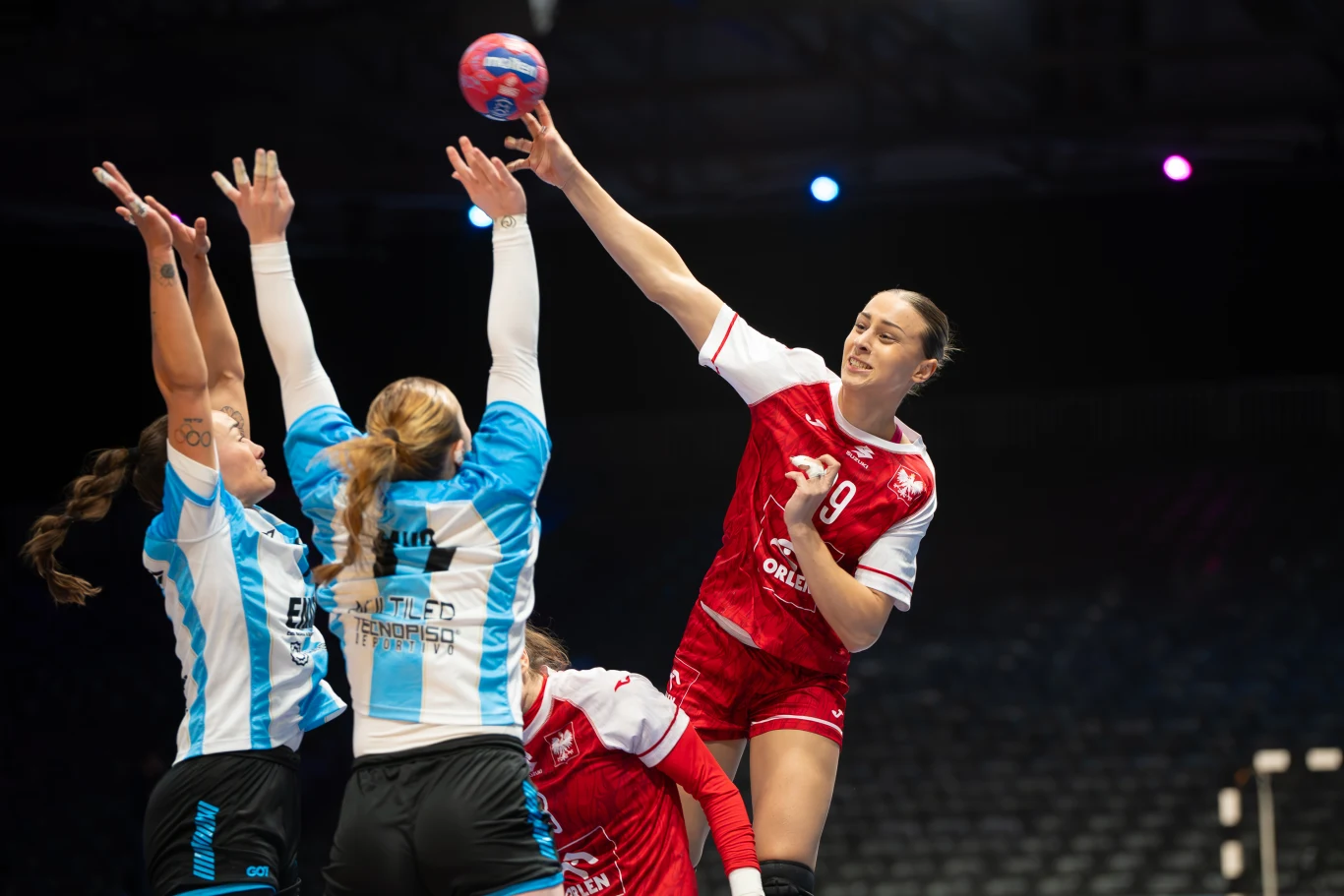 Zawodniczka w czerwonym stroju oddaje rzut piłką ręczną nad blokującymi ją przeciwniczkami w niebiesko-białych koszulkach, akcja rozgrywa się podczas meczu na arenie sportowej.