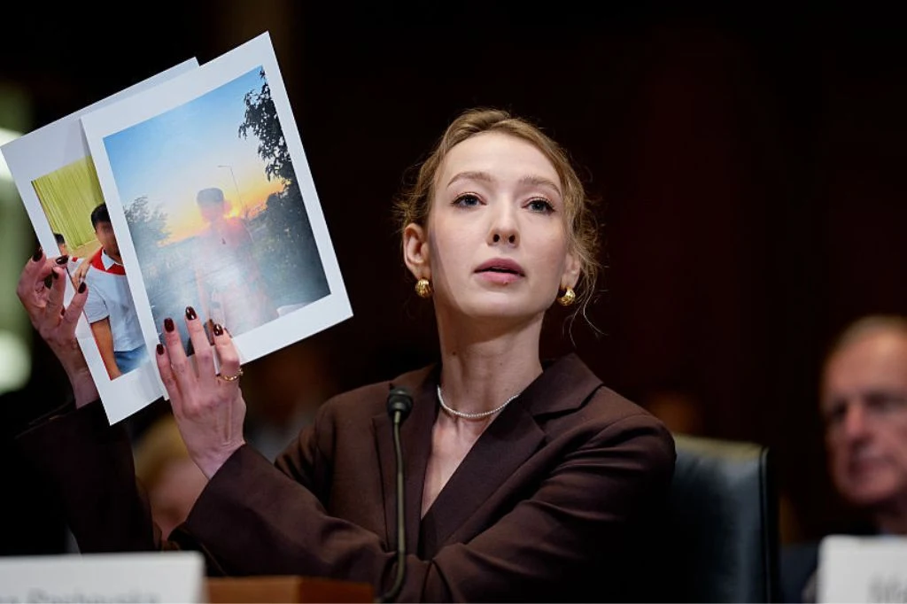 Ekspertka ukraińskiego Regionalnego Centrum Praw Człowieka Kateryna Raszewska Kobieta w garniturze trzyma dwie duże fotografie, wyraźnie prezentując je zgromadzonym. Siedzi za stołem z mikrofonem, w tle widoczne sylwetki innych uczestników spotkania.