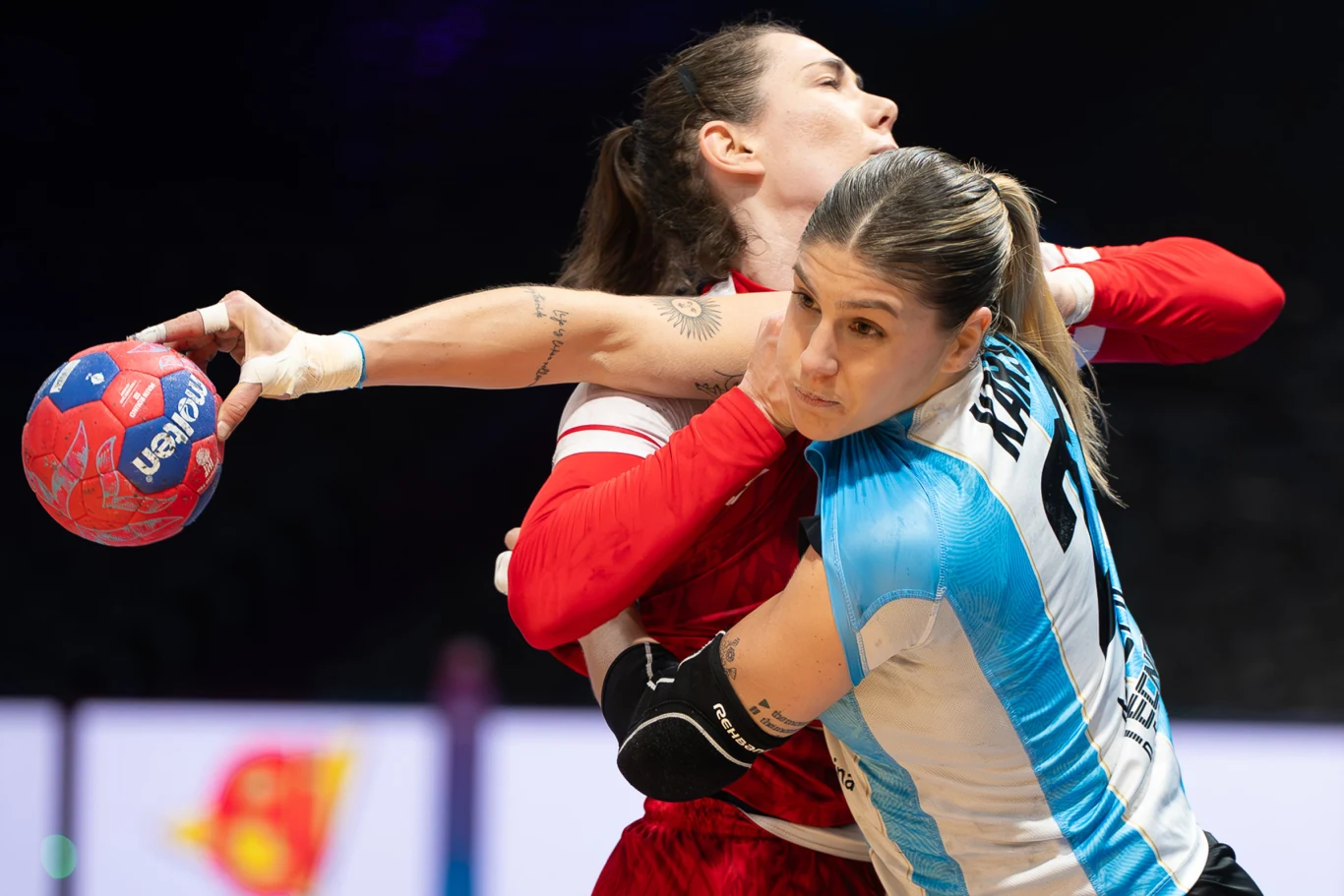 Dwie zawodniczki rywalizujące o piłkę podczas meczu piłki ręcznej, jedna z nich w niebiesko-białej koszulce energicznie blokuje przeciwniczkę w czerwonym stroju.