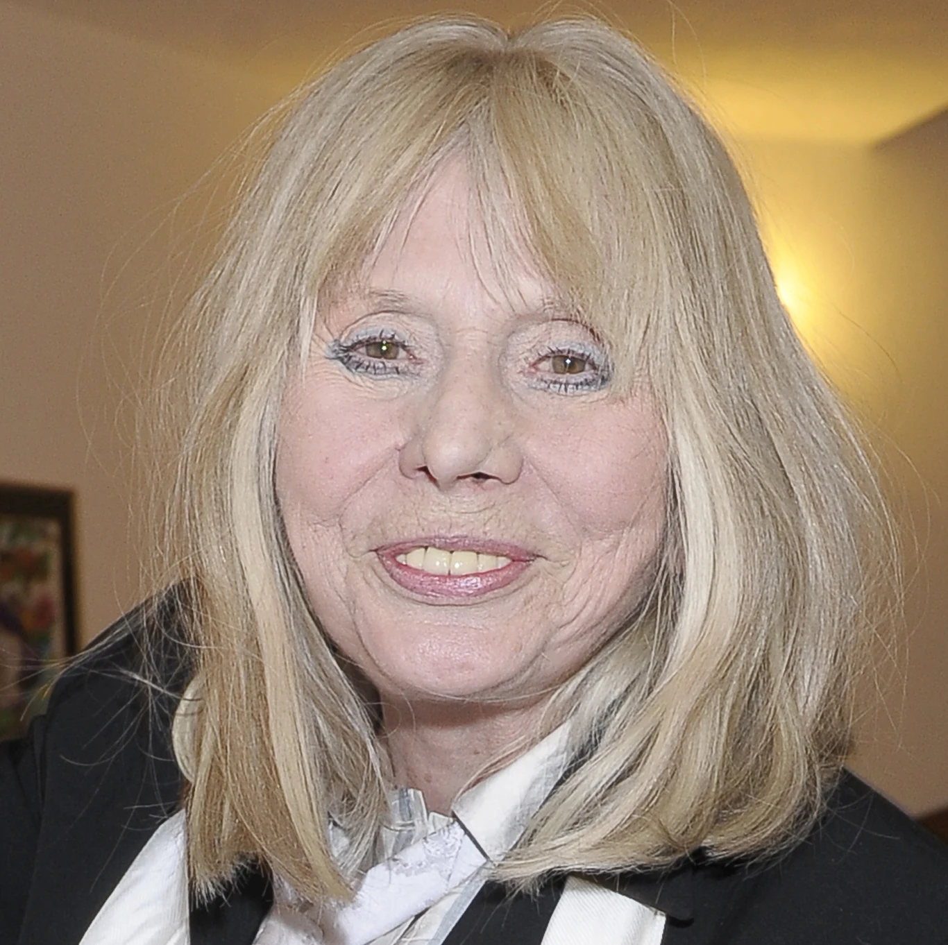 Barbara Wrzesińska Uśmiechnięta kobieta o jasnych blond włosach, ubrana elegancko w biało-czarną koszulę i marynarkę, stoi w ciepło oświetlonym wnętrzu. W tle widoczny fragment ramki ze zdjęciem na ścianie.