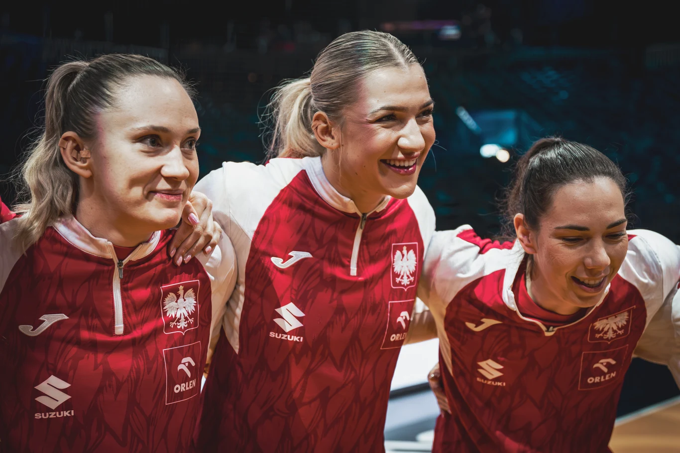 Trzy zawodniczki polskiej reprezentacji w sportowych czerwono-białych strojach uśmiechają się, stojąc ramię w ramię na arenie sportowej.