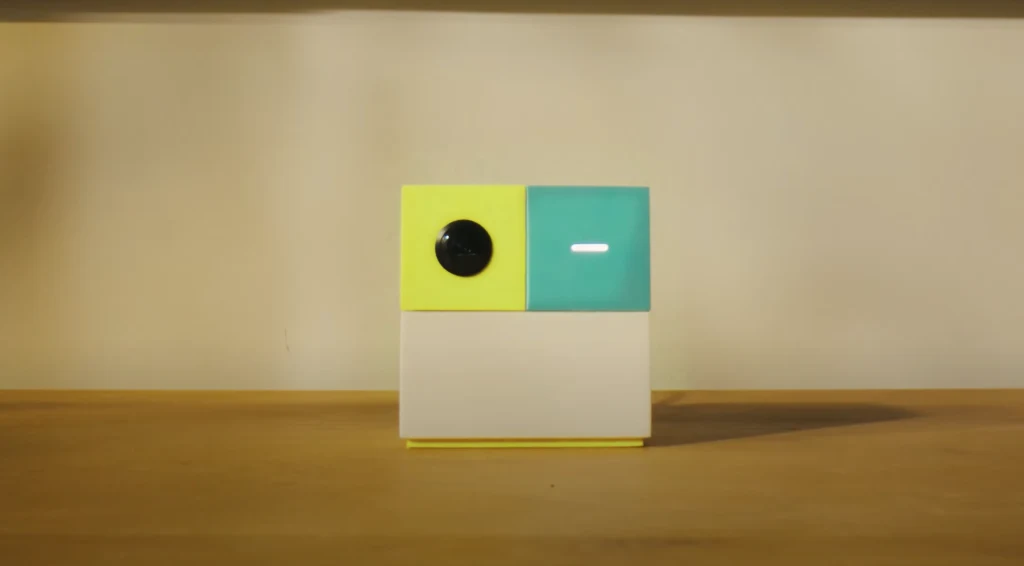 NEX Playground Kolorowy, nowoczesny gadżet o geometrycznym kształcie, składający się z dwóch górnych sześcianów – żółtego z okrągłym czarnym elementem i niebieskiego z poziomym białym paskiem – umieszczonych na białej podstawie, ustawiony na drewnianej powierzchni na...