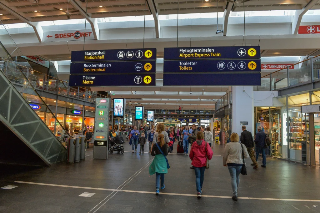 Zainteresowanie pracą i mieszkaniem w Norwegii wśród Polaków spadło Wnętrze nowoczesnego dworca kolejowego z tłumem ludzi, wyraźnymi tablicami informacyjnymi z oznaczeniami kierunków oraz sklepami i lokalami usługowymi po bokach, wokół panuje codzienny ruch.