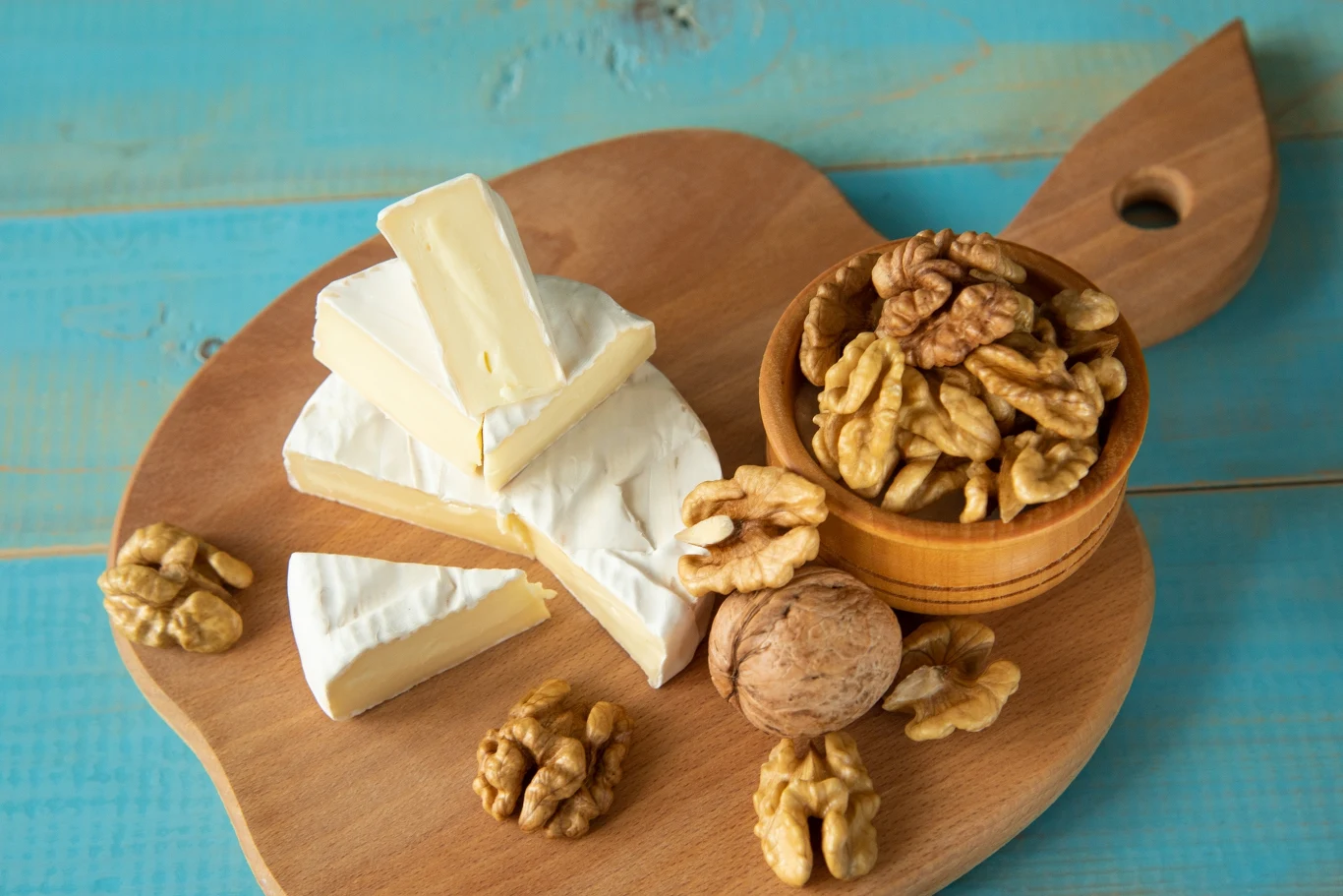 Do przygotowania świątecznej przekąski będziesz potrzebować między innymi sera camembert i orzechów Kawałki białego sera pleśniowego oraz orzechy włoskie, część w miseczce, część rozrzucona na drewnianej desce w kształcie palety, ustawione na niebieskim drewnianym tle.