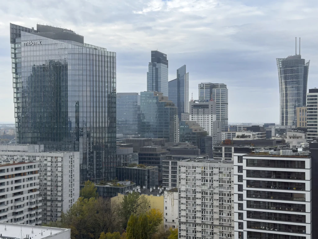 Polacy źle ocenią sytuację finansową kraju? "Tradycyjnie lubią narzekać" Nowoczesne wieżowce ze szkła i betonu w centrum miasta, dominujące nad niższymi blokami mieszkalnymi, z widocznym logo banku na jednym z budynków oraz szarym, pochmurnym niebem w tle.