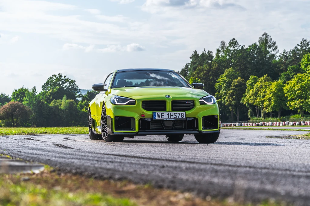 Jaskrawożółty samochód BMW M2 poruszający się po asfaltowym torze z widocznym śladem po ślizgu na zakręcie, w tle drzewa i fragment nieba z rozproszonymi chmurami.