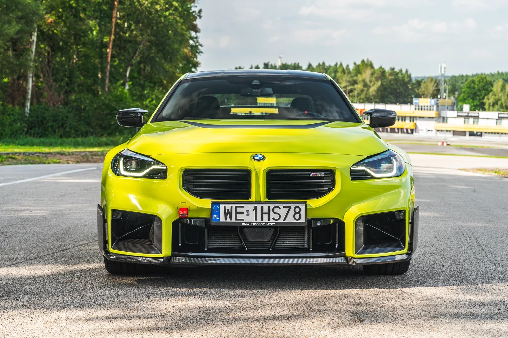 Limonkowy sportowy samochód marki BMW na środku pustej asfaltowej drogi, otoczony zielenią drzew oraz z widoczną tablicą rejestracyjną z przodu.