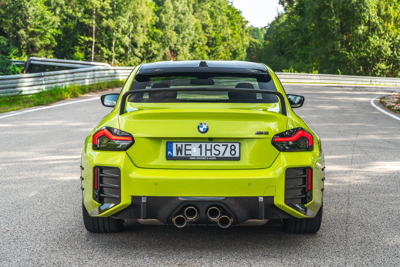 BMW M2 Jaskrawozielony sportowy samochód BMW M4 z widocznym wyraźnie tyłem oraz czterema rurami wydechowymi stoi na zakręcie asfaltowej drogi otoczonej lasem, z polską tablicą rejestracyjną.